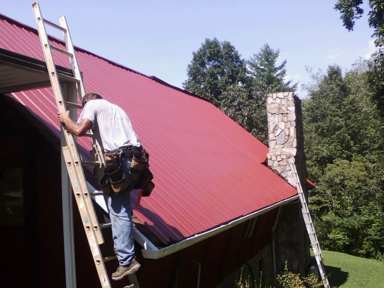 Roofer on a ladder beside a red metal roof and stone chimney — Soffit & Fascia Repair by Roofing Brothers in Newtonville